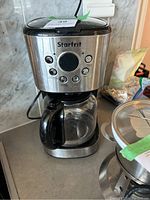 Front angled photo of the Starfrit coffee machine showing stainless steel body, control buttons, and included glass carafe with black handle.