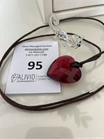 Swarovski cat figurine placed next to a red heart pendant necklace on a brown cord with auction tag visible.