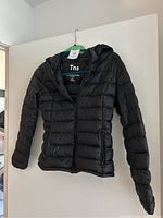 Black Tna puffer jacket hanging on green hanger showing front and hood