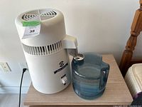 Side view of electric water distiller sitting next to blue glass jug on wooden surface, distiller powered by cord.
