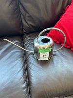 Full view of the silver metal watering canister placed on a dark leather surface with a red cushion in the background, showing its shape including the long spout and circular handle.