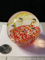 Side view of the large glass paperweight showing the reddish-orange and white streaked center with yellow swirls and embedded glass bubbles, quarter for scale.