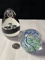 Oblong tapered clear glass paperweight with internal bubble design beside a quarter for size reference, with round globe paperweight featuring blue and green swirls.