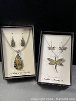 Two jewelry gift sets in black boxes: one with teardrop pendant and earrings featuring greenish-yellow stones and silver swirl accents, the other with dragonfly pendant and earrings with yellow-green accents.