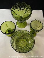 Four olive green depression glass items shown on white background, including a pedestal bowl, two goblets, and a divided dish