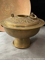 Side view of the antique brass rice cooker on a pedestal base with detailed zodiac animal motifs on the lid, one handle present, the other missing.