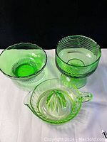 Three green glassware items shown under normal light: a fluted bowl, a footed swirl-patterned compote, and a lemon juicer with pouring spout and reamer.