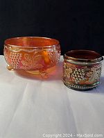 Front angle view of both orange and purple grape harvest themed carnival glass pieces.