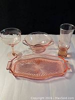 Four pink Depression glassware pieces: a coupe cocktail glass, two-handled bowl, tall narrow vase, and rectangular serving tray with pinwheel design.