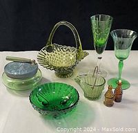 Lot of green glassware items arranged on a white surface including basket, bowls, two stemmed glasses, a lidded jar with atomizer, and two miniature brown bottles.