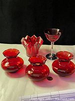 Five ruby red glass items arranged on white surface with black background. Three small round vases in front row; behind them a star-shaped sawtooth edge dish and a tall pedestal glass.