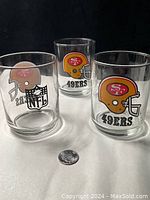 Three vintage NFL 49ers bucket style glasses displayed on black background with a quarter for scale. Two have gold 49ers helmet logos, one has brown helmet NFL logo.