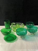 Seven green glassware items arranged side by side on black background including lemon juicer, bowls, glasses, and dessert dishes
