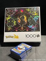Photo of the 1000-piece Pokémon puzzle box with black silhouette Pokémon art and a stack of Pokémon trading cards in front.