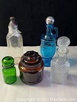 Five lidded glass bottles and jar: green, clear Pyrex, amber jar, cobalt blue, clear decanter.