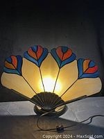 Front view of the vintage stained glass peacock feather table lamp illuminated, showing the multicolored fan-shaped shade.
