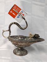 Side angle view showing detailed floral embossing, curved handle, wick setup with cylindrical holder, and overall silver patina.