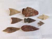 Top-down view of seven Indigenous-style stone arrowheads and spearheads arranged on a white cloth background. Various shapes and colors visible.