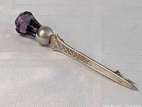 Large sterling silver Scottish clan pin laid on white textured fabric, showing detailed Celtic knot work on the stem and large faceted amethyst at top.