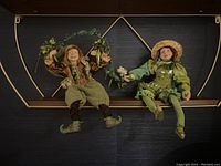 Two elf figurines seated on a wooden shelf with a gold geometric metal frame, one holding a leaf garland, the other holding flowers.