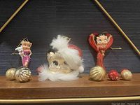 Three Betty Boop Christmas ornaments, two standing and one face with Santa hat, displayed with small Christmas ball ornaments in gold and red.