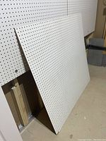 White peg board panel leaning against wall, showing full surface with evenly spaced holes.