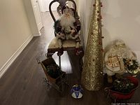 Photo showing gold cone-shaped Christmas tree, two Santa Claus figures, one seated on a chair and one standing, two slate sled decorations with faux pine and pinecones, and other assorted Christmas ornaments including a blue and white ornament and red sleigh bowl.