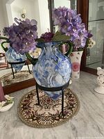 Blue and white art glass vase with green handles on black metal stand on a decorative circular placemat, photographed alongside a mirror and other decorative items.