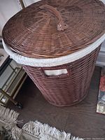 Wicker laundry basket with dark brown woven rattan, fabric lining, lid, and side handle on hardwood floor next to a small table.