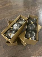 Two gold boxes displaying eight new clear glass Christmas ornaments with decorative images inside, arranged in protective slots.