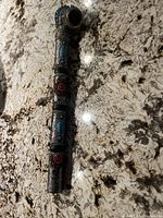 Decorative metal pipe standing vertically on a granite countertop highlighting turquoise and red stone inlays and detailed metal texture.