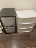Two plastic drawer storage units side by side on wooden floor, white with three drawers and black with two drawers.