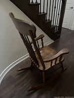 Wooden rocking chair positioned angled left showing its curved seat, armrests and rockers on dark flooring.