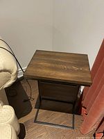 Wooden speaker table with dark brown wood grain top and metal frame base shown near furniture and wall outlet.