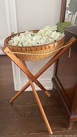 Front view of wooden X-frame stand with woven basket filled with white artificial hydrangea flowers, placed indoors on wood floor.