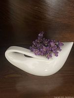 Front angle view of white porcelain swan vase with purple flowers inside, showing smooth glossy finish and elegant design.