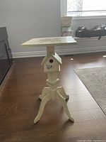 Side view of wooden side table showing birdhouse detail and curved legs on hardwood floor.