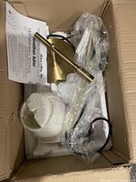 Unopened wall sconce components including a gold metal conical base and cylindrical lamp holder wrapped in plastic inside cardboard box with packing.
