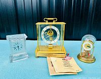 Wide shot showing all three clocks placed on a blue surface against a dark background: crystal rectangular clock, brass carriage clock, and small clock under glass dome.