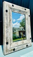 Angled view of farmhouse rustic mirror showing distressed white wooden frame with wrought iron bolt accents and reflective glass surface.