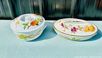 Two Royal Worcester covered serving dishes, one round and one oval, on a light blue cloth background showing their Evesham fruit and wheat design on lids and bodies.