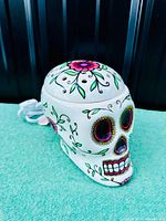 Front angle photo of the white ceramic sugar skull-shaped wax warmer lamp with colorful hand-painted floral and vine designs, showing powered cord and intricate details on the skull face.