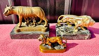 Front view of three vintage cast metal animal sculptures on reddish fabric backdrop. Includes Romulus and Remus she-wolf and two lion sculptures, all on marble or solid bases.