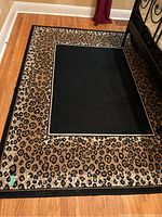 Full view of rectangular area rug with leopard print border and solid black center panel on a hardwood floor under bed frame.