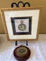 Photo of entire lot showing round vintage barometer on table and gold framed wall art with pocket watch image on black and script background.