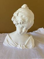 Front view of white ceramic bust depicting a young girl with downward gaze, detailed facial features and hair texture.