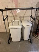 Front view of adjustable dual clothes rack with two white bins beneath it, one rectangular with vented sides and one tall oval can.