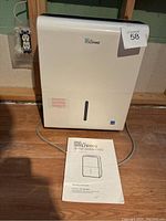 Full front view of the white Living dehumidifier with control panel visible and a small water window at front, placed on a wood floor near an electrical outlet. Assembly instructions manual placed in front.