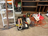 Photo showing white metallic stepladder, pick ax with wooden handle, black claw hammer, assorted hand tools, and orange extension cord bundled coiled on floor