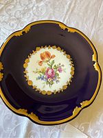 Top view of round decorative plate with kobalt blue background, gold decorative rim, and floral painted center bouquet.